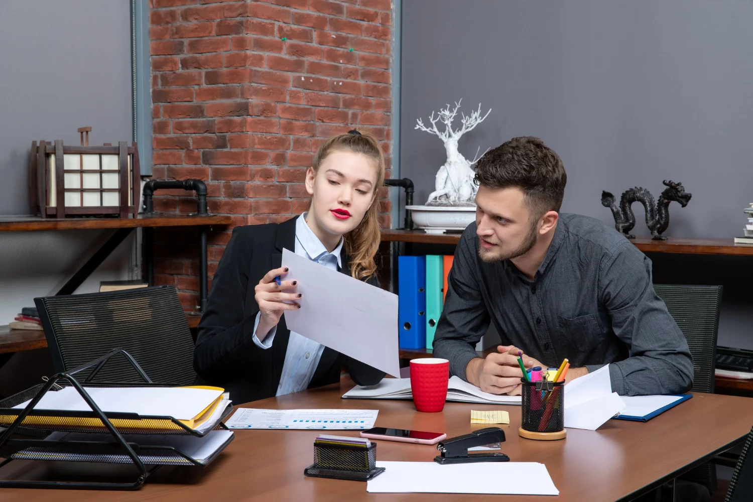 Two professionals discussing documents in a modern office setting with stationery and a coffee cup on the table.