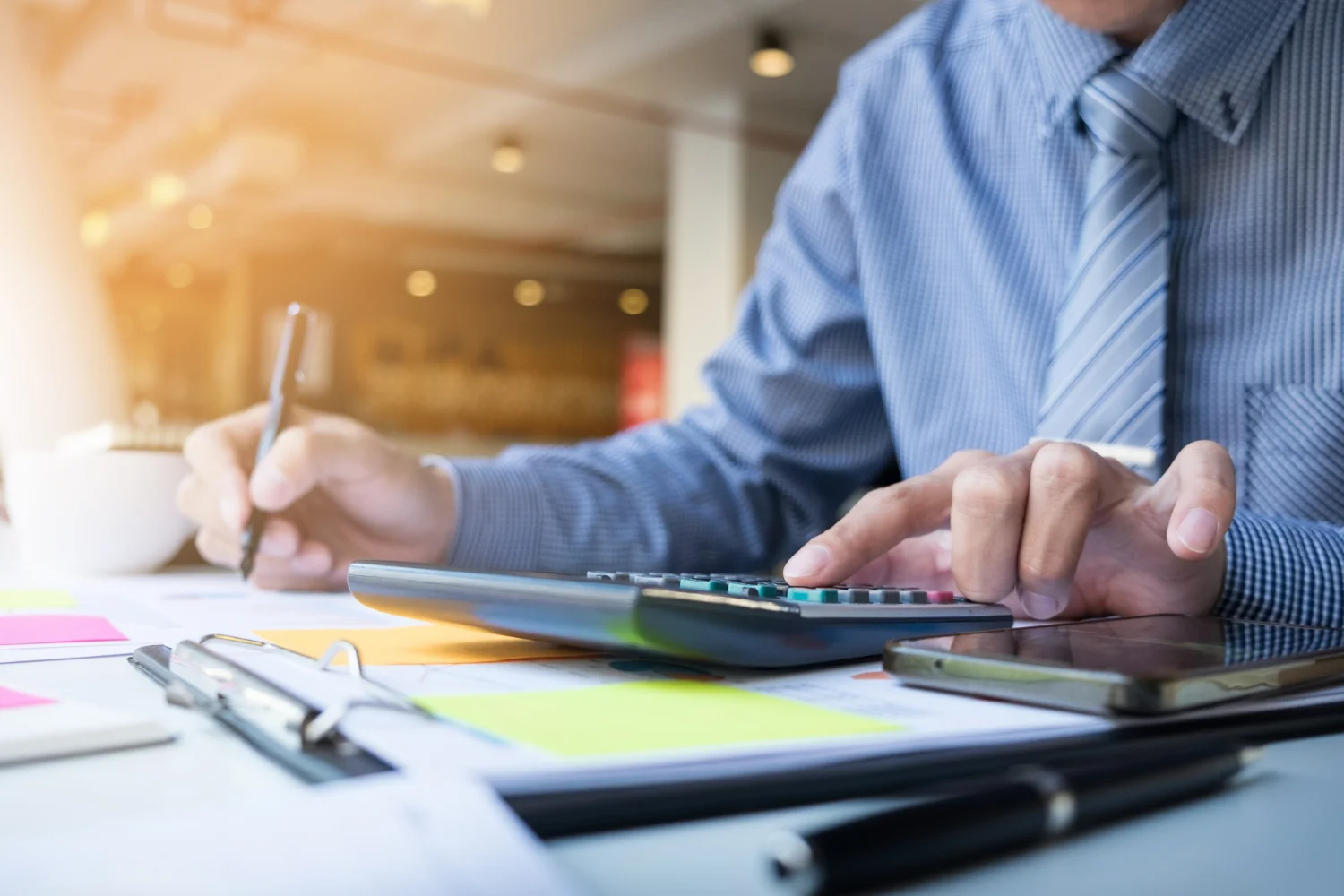 A person in a striped shirt diligently tackles accounting tasks at a desk, skillfully using a calculator amidst scattered documents, a smartphone, and a cup.