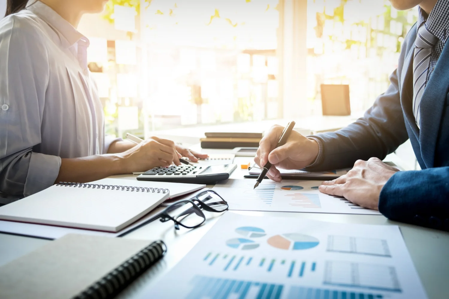 Two professionals analyzing financial data with a calculator, charts, and graphs on a desk during a business meeting.