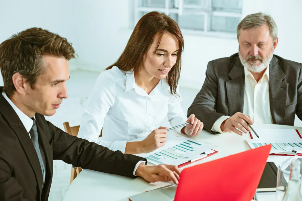 Three professionals engaged in a business meeting, analyzing data on a laptop and discussing reports with charts and graphs.