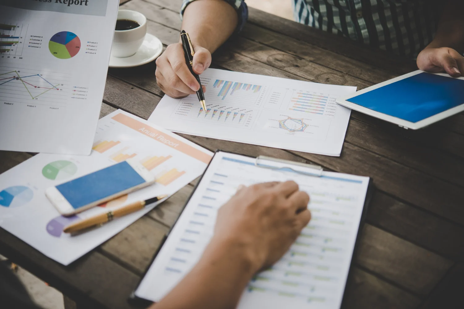 Two individuals analyzing business reports with charts, graphs, and a tablet on a wooden table, showcasing data analysis and collaboration.