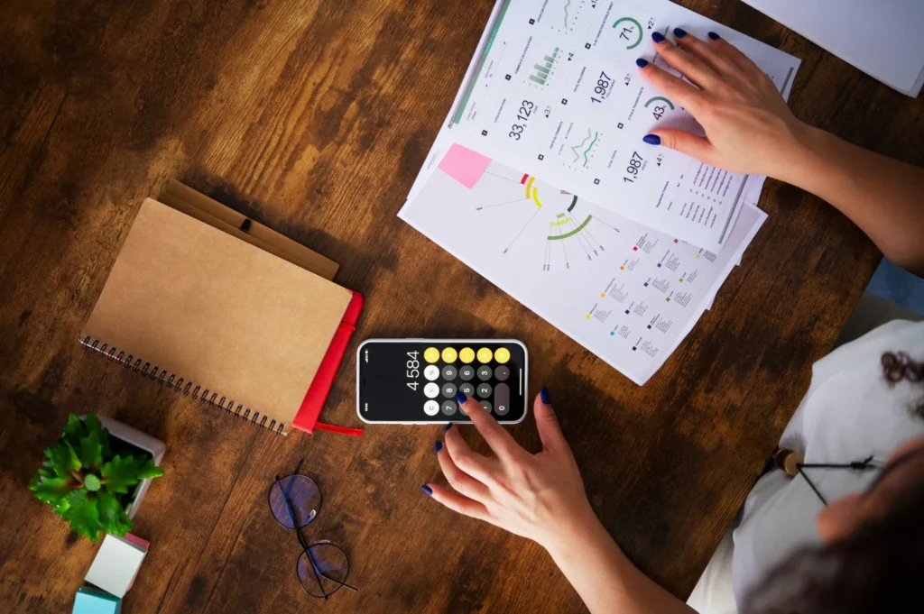 A person using a calculator on a smartphone while reviewing business reports and charts on a wooden table with a notebook and glasses.