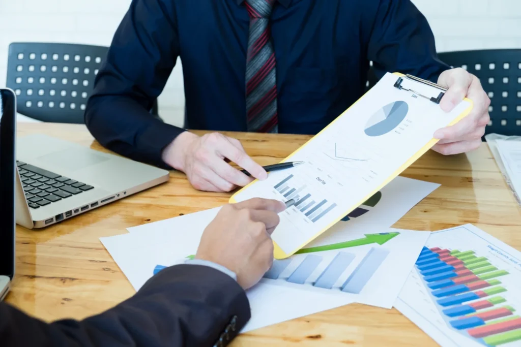 Two professionals discussing business reports with charts and graphs on a table, including a laptop and documents.