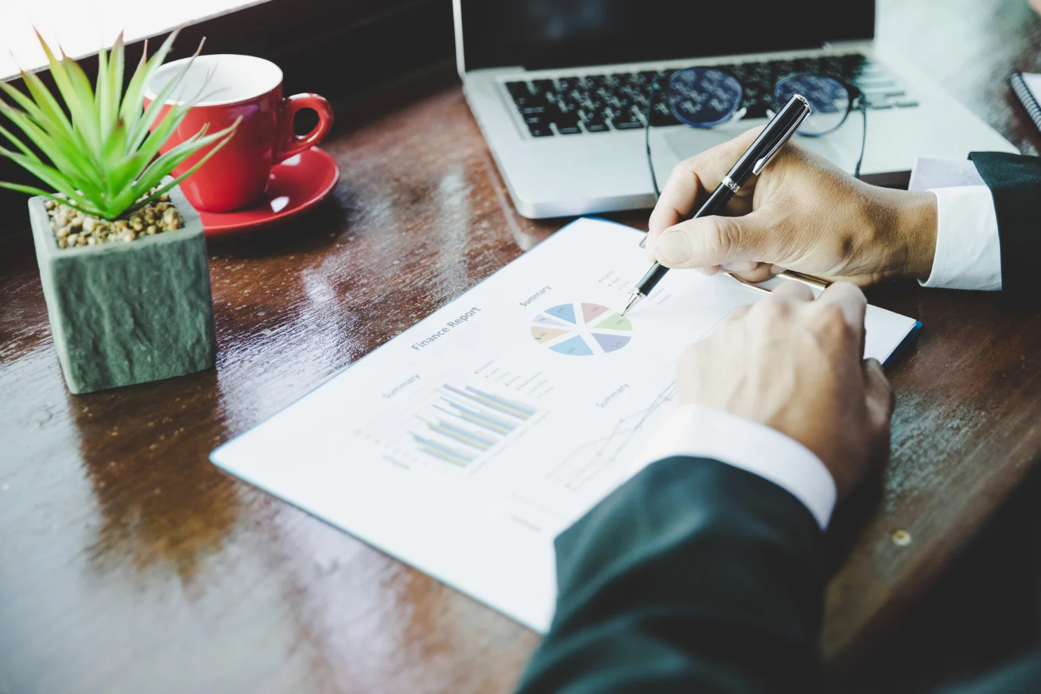 A person analyzing a financial report with charts and graphs, using a pen, alongside a laptop and a cup of coffee.