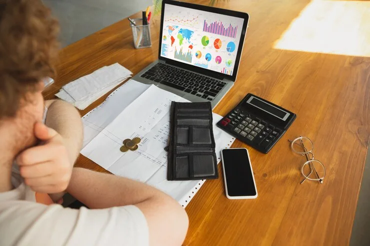 A workspace featuring a laptop displaying financial graphs, a wallet, coins, a calculator, and a smartphone on a wooden table.