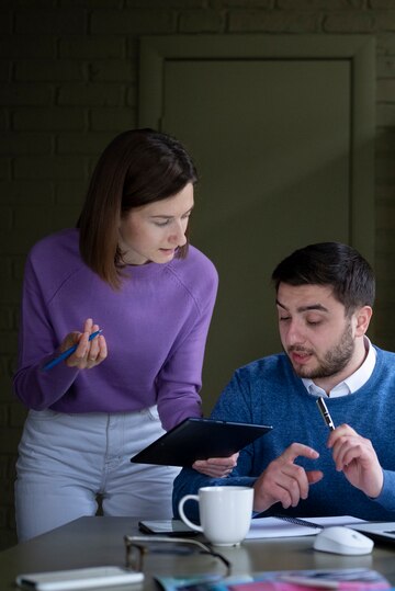 A woman in a purple sweater shows a tablet to a man in a blue sweater during a business meeting, with a coffee cup on the table.